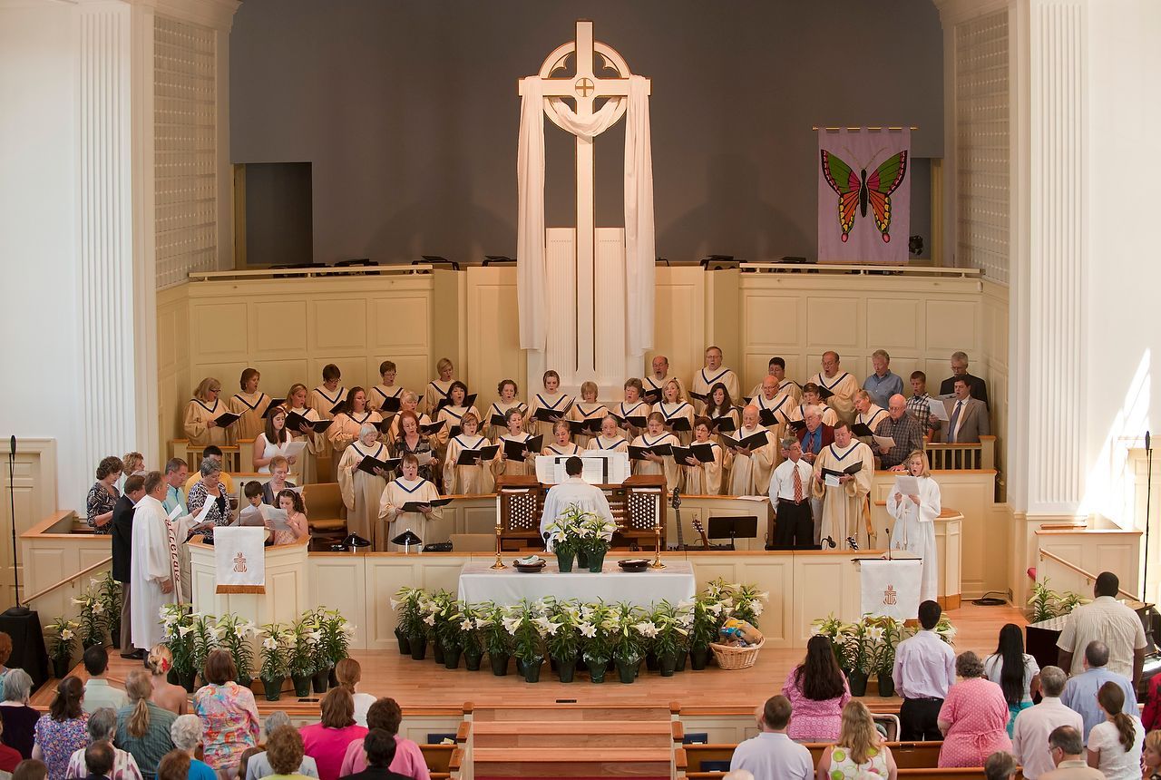 Choir performing on stage in a church. People in pews watch. White flowers decorate the stage.