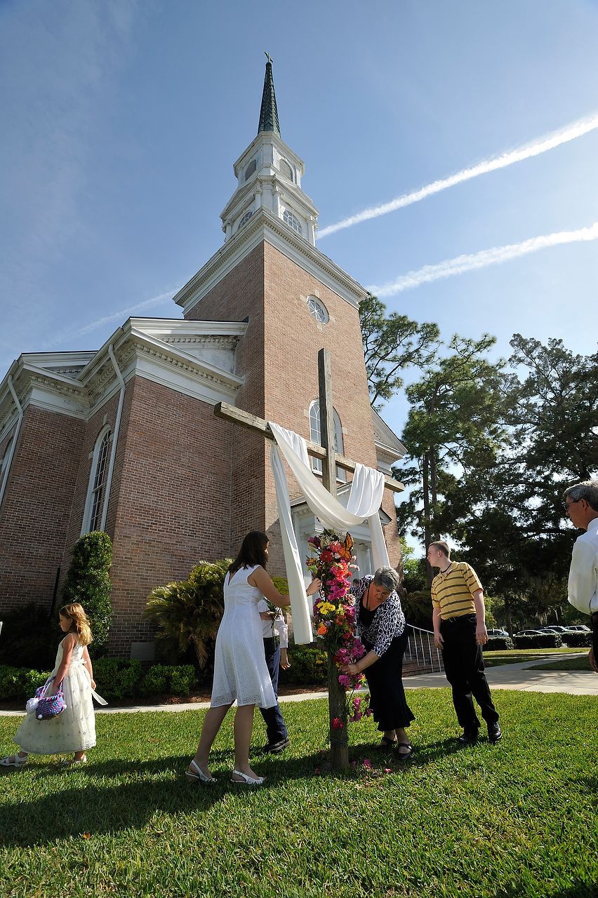 People decorating a cross with flowers outdoors, church building in the background. Bright sunny day.