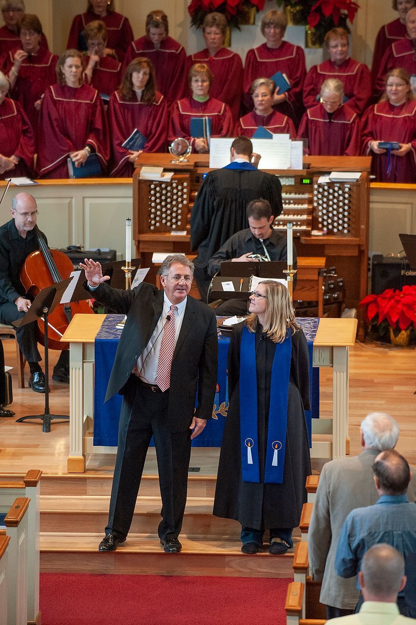 Choir and musicians in church; conductor gestures, woman in robes stands by.