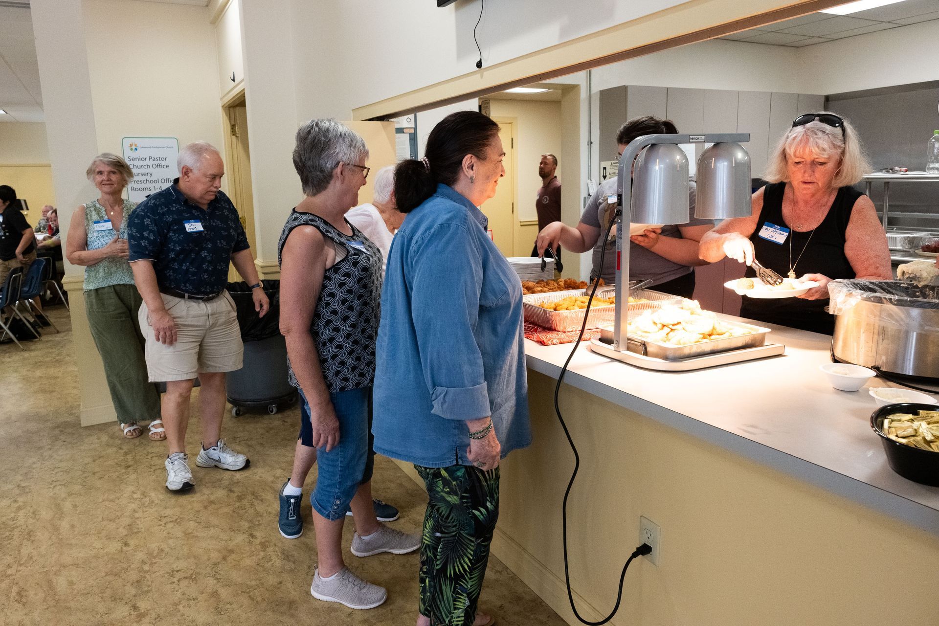 People line up at a buffet, served by a woman in a black shirt.