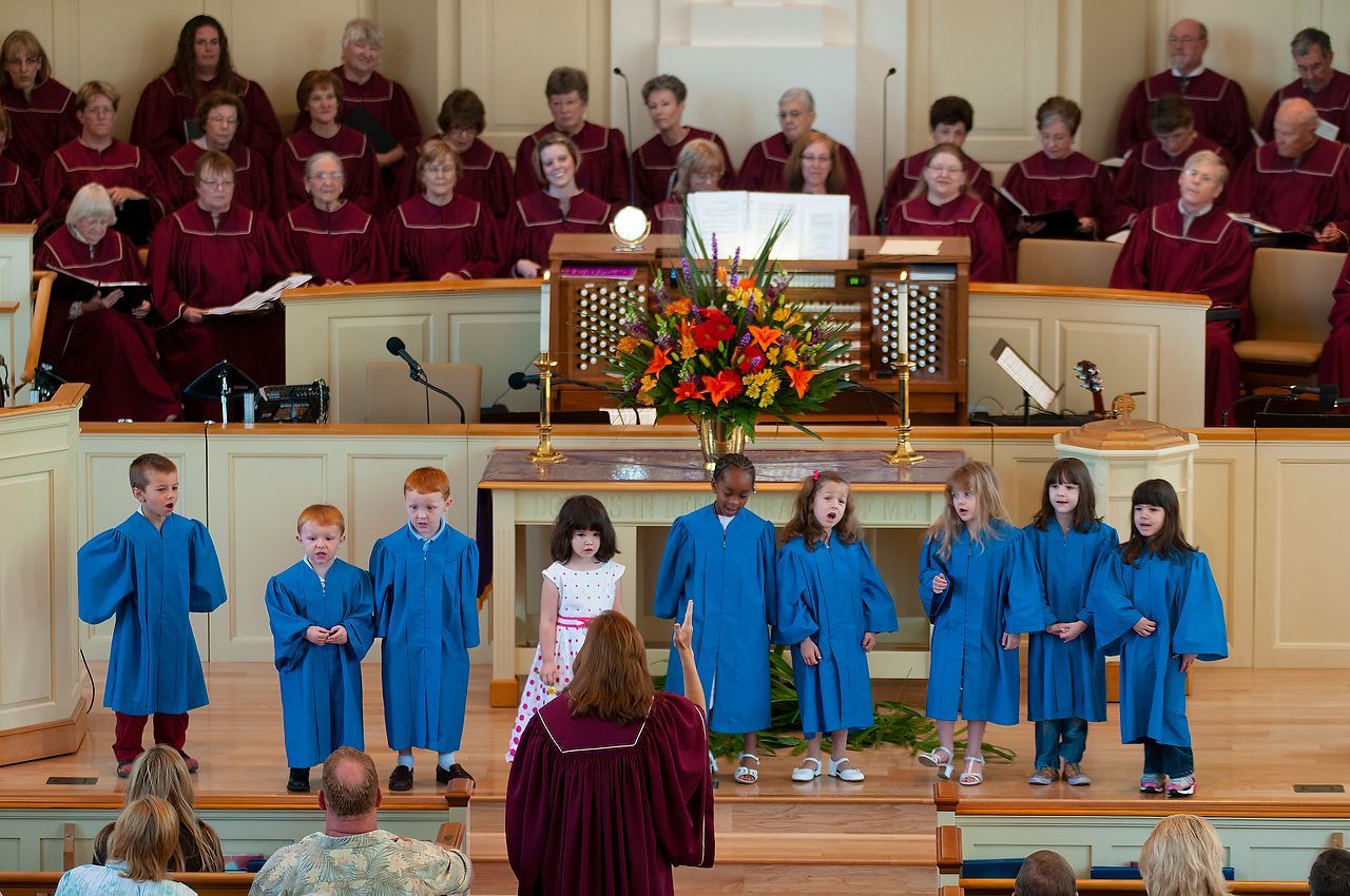 Children and choir in robes singing on a stage, with an organ, floral arrangement, and conductor in a church.