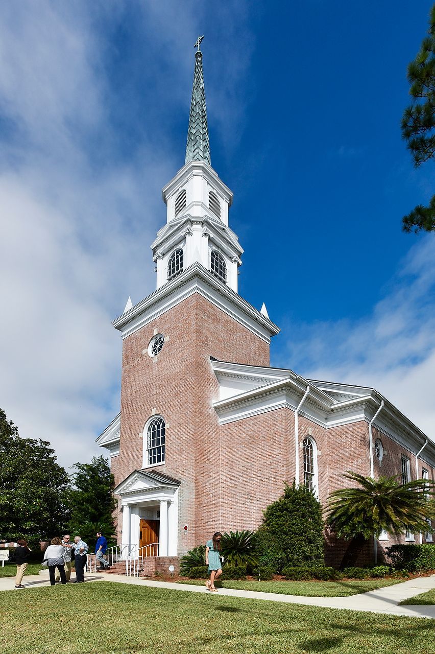 Brick church with white trim and steeple, people walking on the lawn under a blue sky.
