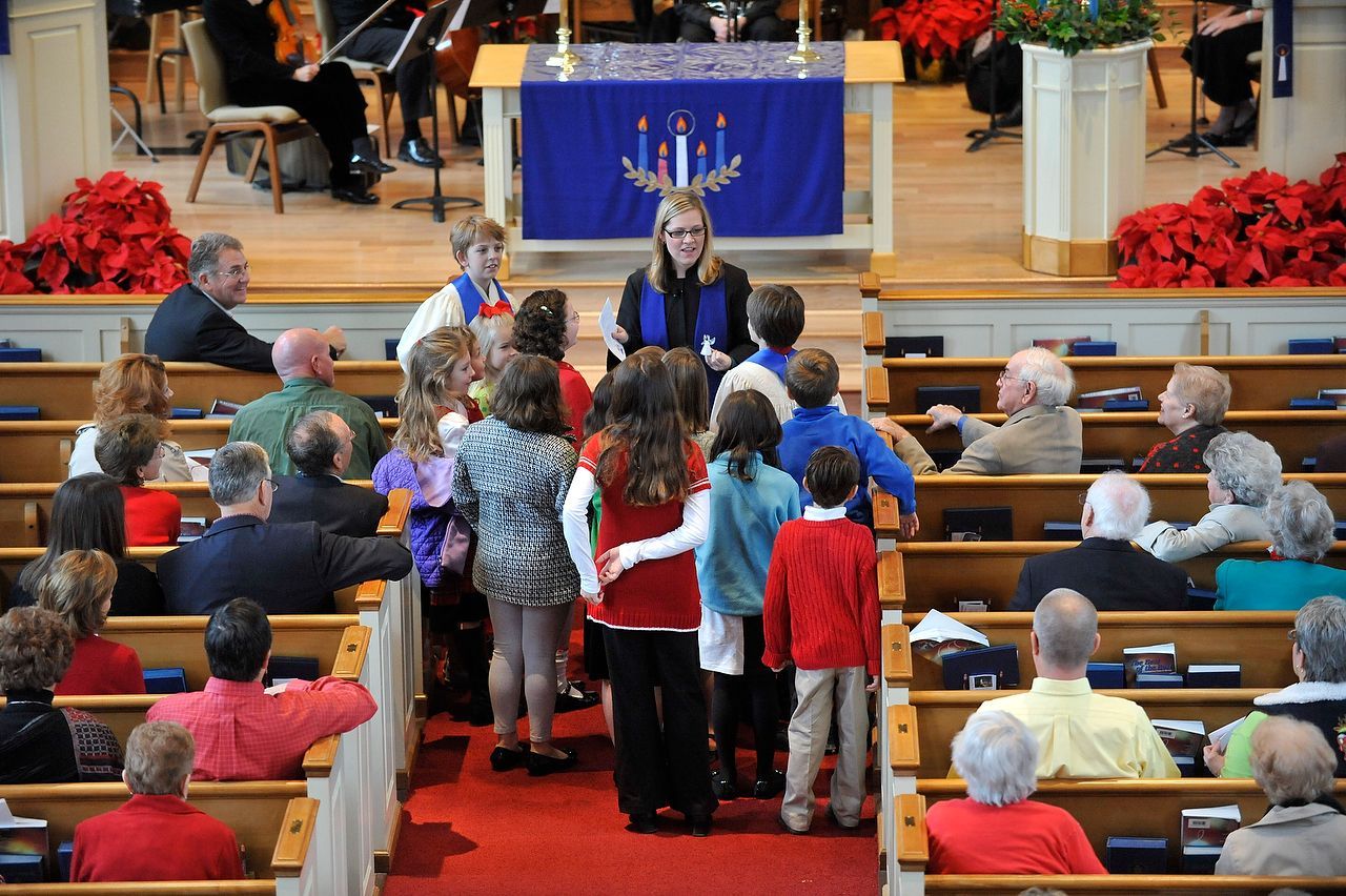 People in a church listen to a woman speaking from a podium. Children walk up the aisle.