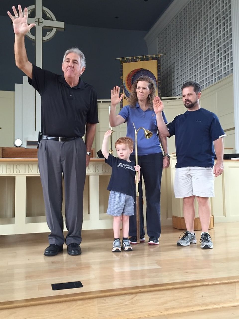 Four people standing on a stage, raising their hands. A child holds a brass instrument. Church interior.