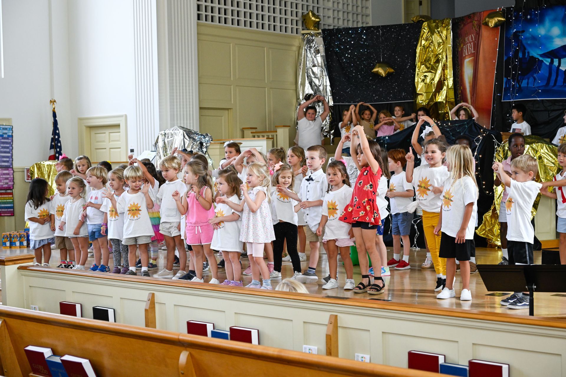 Children in white shirts and various clothes on a stage, some raising their hands, in front of a colorful backdrop.