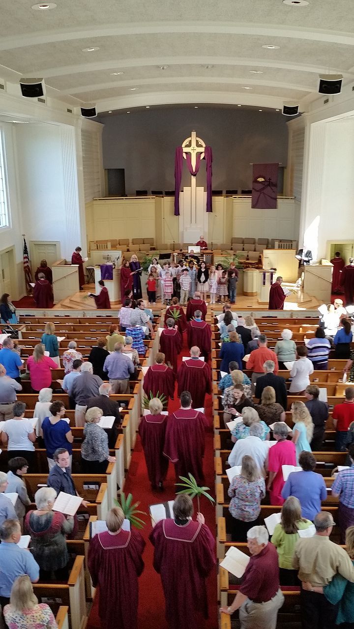 Church service in progress: people seated in pews, clergy in maroon robes walking toward altar.