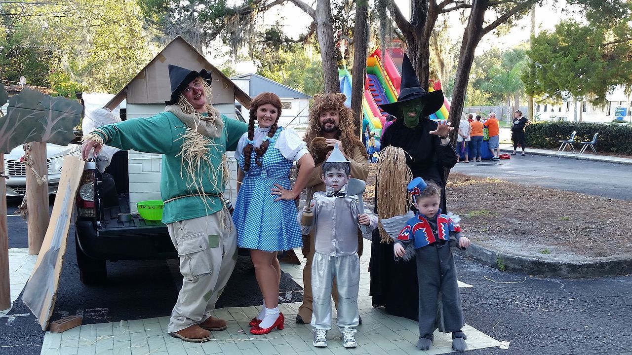 People in Wizard of Oz costumes pose outside: Scarecrow, Dorothy, Cowardly Lion, Tin Man, Wicked Witch, and a Munchkin.