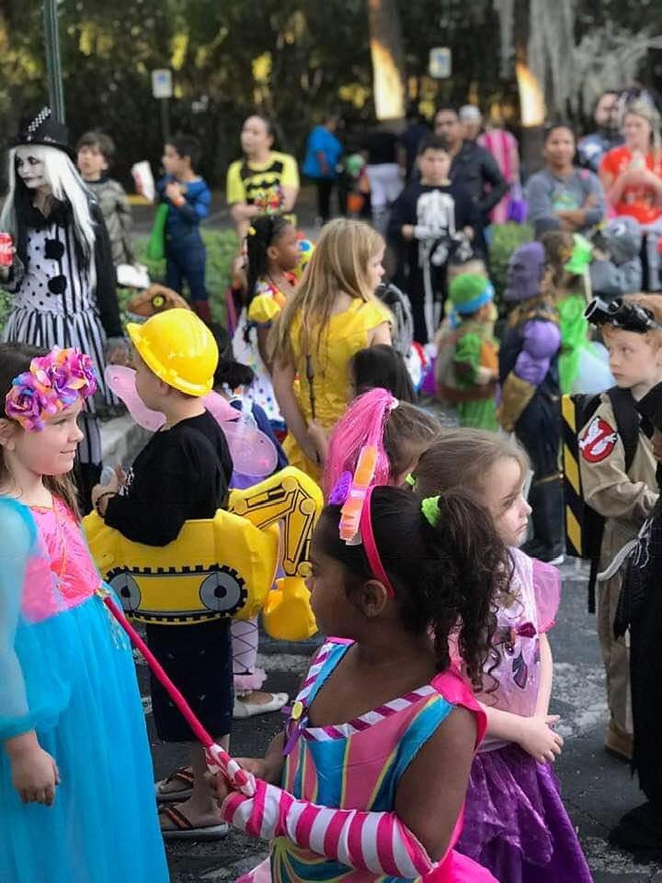 Children in Halloween costumes gathered outdoors, some holding props; others are watching.