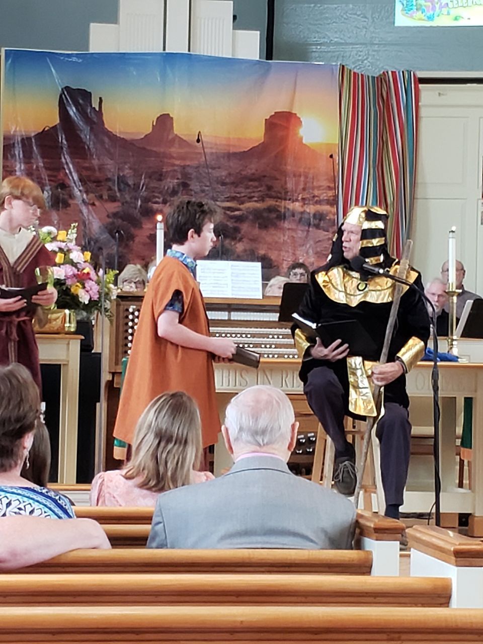 Church play with actors in costume; backdrop of desert sunset; audience seated.