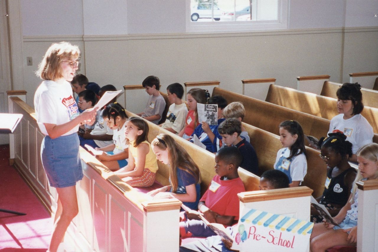 Children in church pews, listening to a woman read from a paper. 
