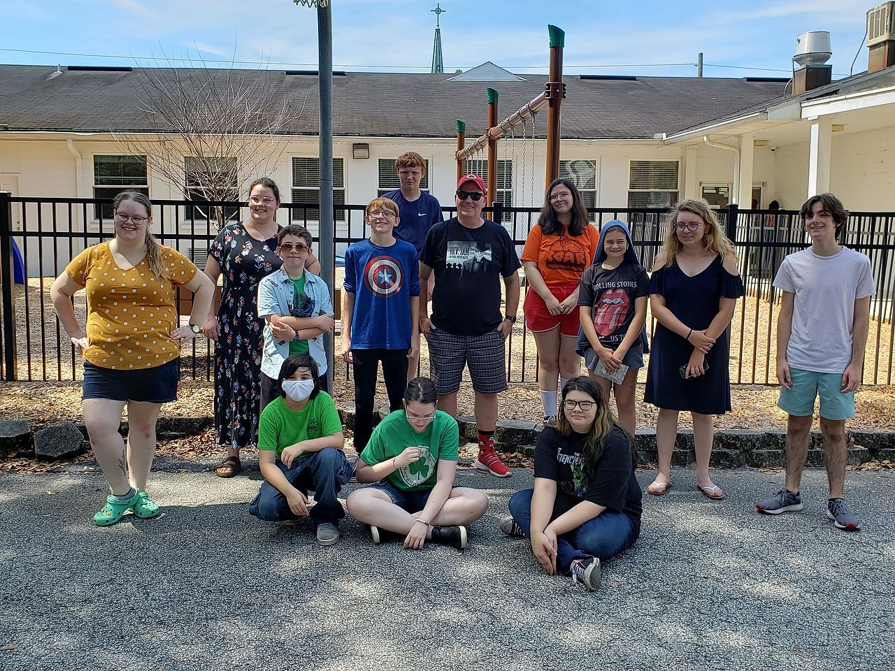 Group of people poses outdoors near a playground. Some stand, others sit. Bright sunny day.