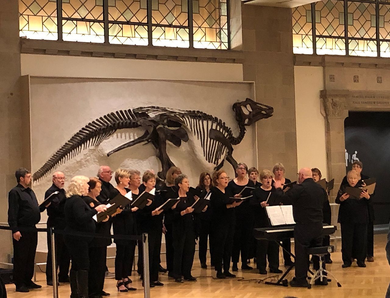 Choir performing in front of a dinosaur skeleton in a museum.