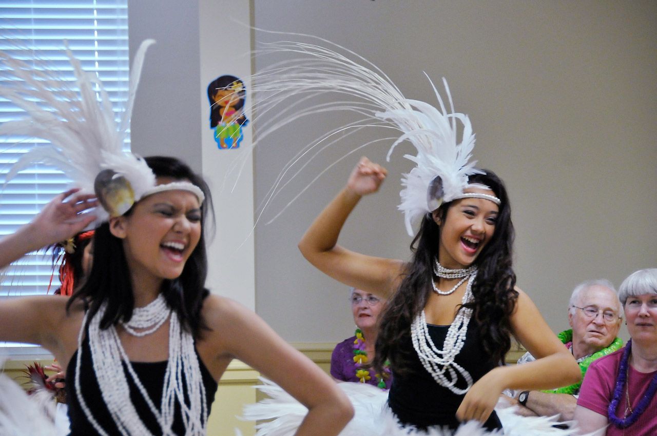 Two dancers in white feather headdresses perform, smiling, in front of an audience.