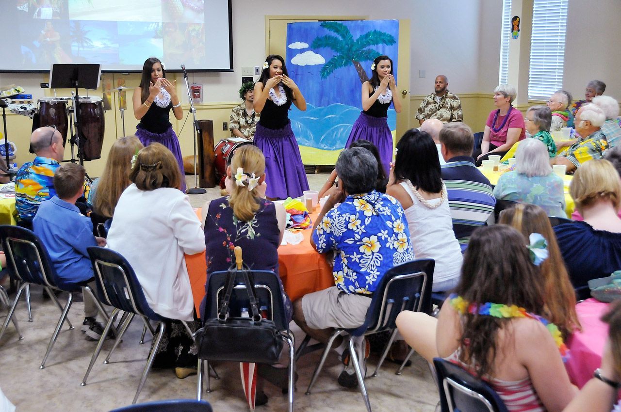 Hula dancers perform at a party with people seated at tables, decorated in tropical colors.