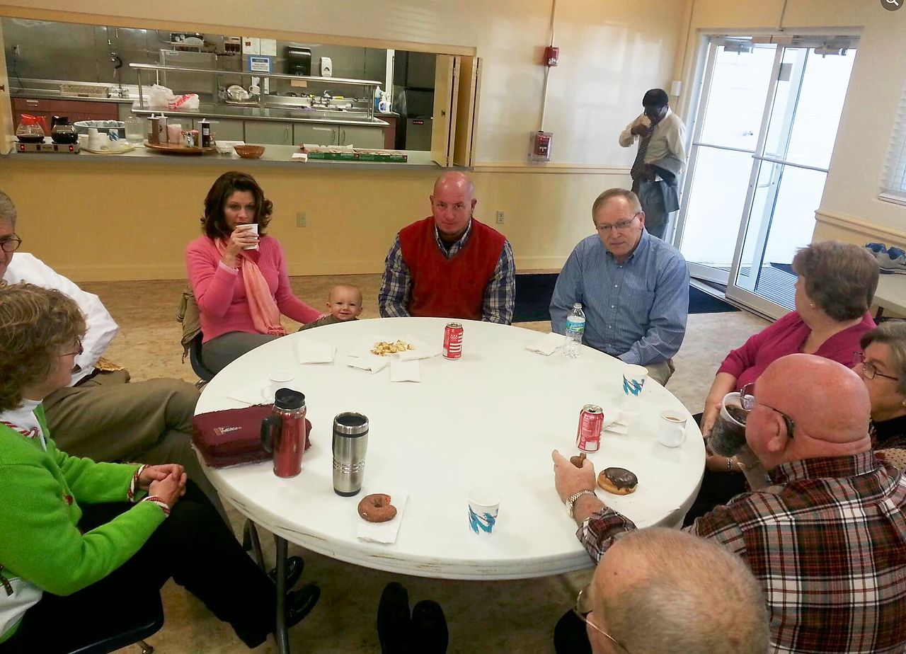 People sit around a white circular table, talking and drinking. Some items are on the table, likely food. Bright setting.