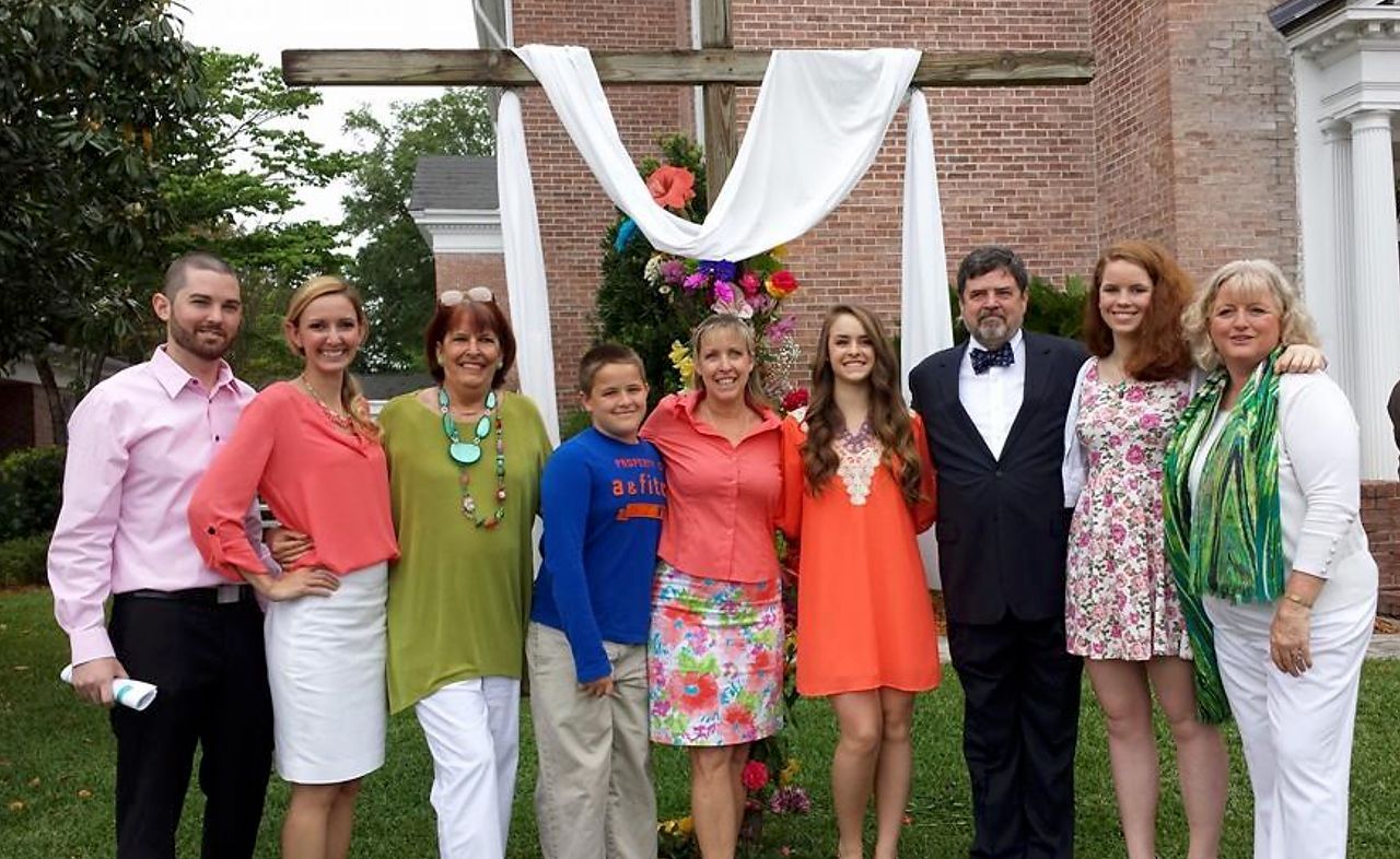 Group of people smiling in front of a draped cross outside a brick building.