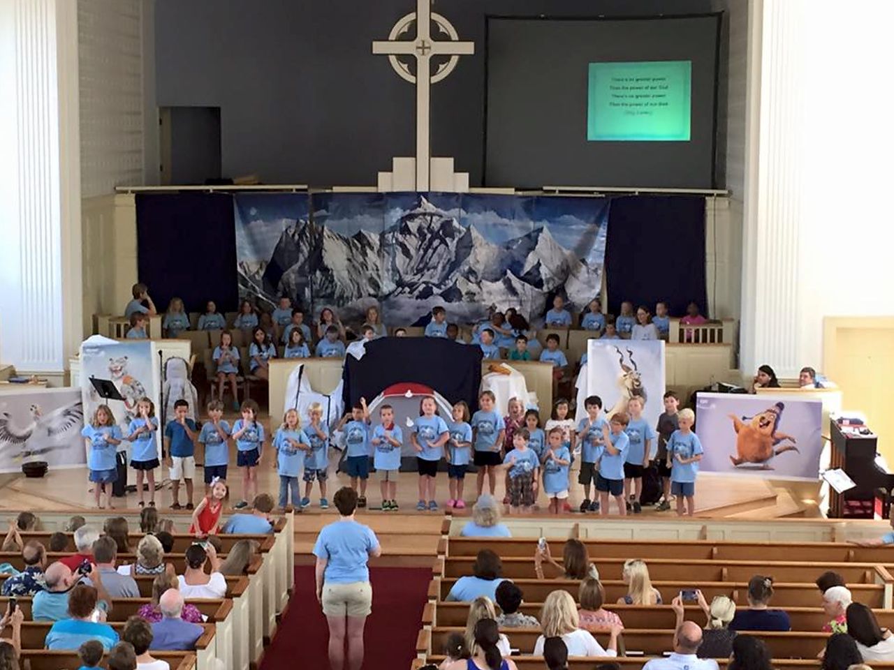 Children performing a play on a church stage; audience watches. Backdrop of mountains, cross above.