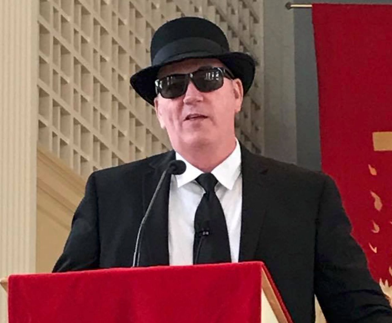Man in a Blues Brothers suit, hat, and sunglasses speaking at a red podium in a church.