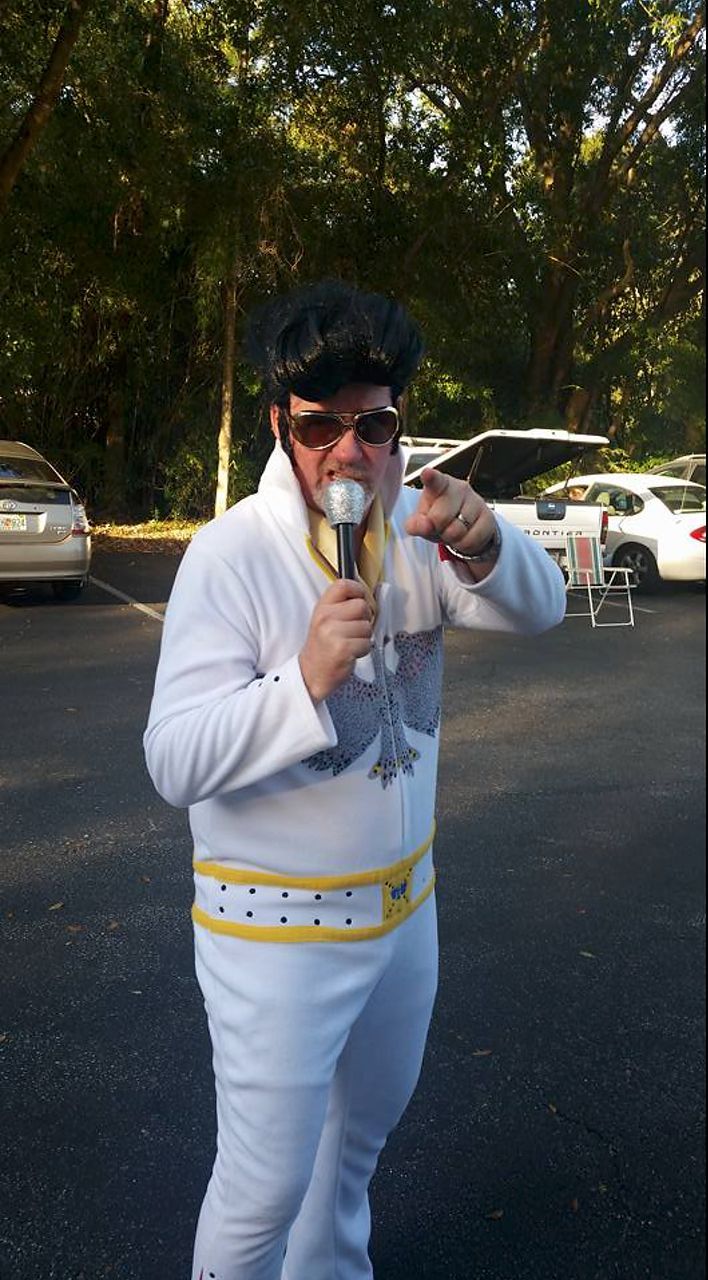 Man dressed as Elvis, in white jumpsuit, holding microphone, pointing, outside in parking lot.