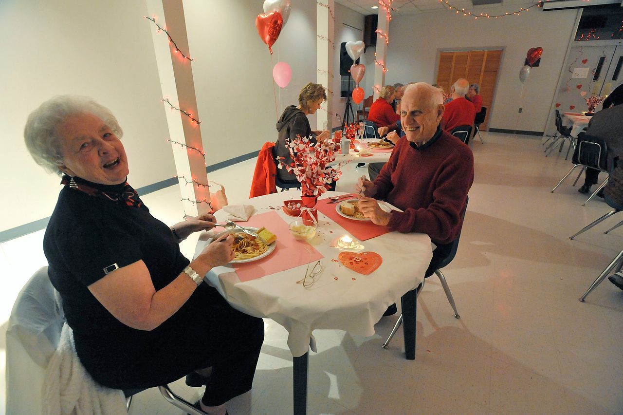 Two people seated at a table, eating at a Valentine's Day event. Decorations include balloons and hearts.