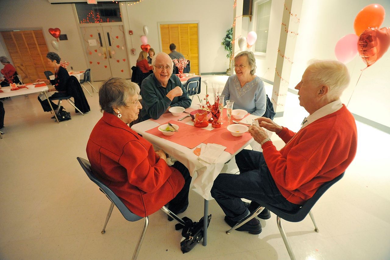 People at tables in a room, some wearing red, with Valentine's Day decorations and balloons.