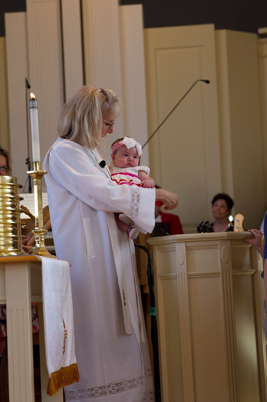 Clergyman in white robes holds a baby with a floral headband during a christening at a church.