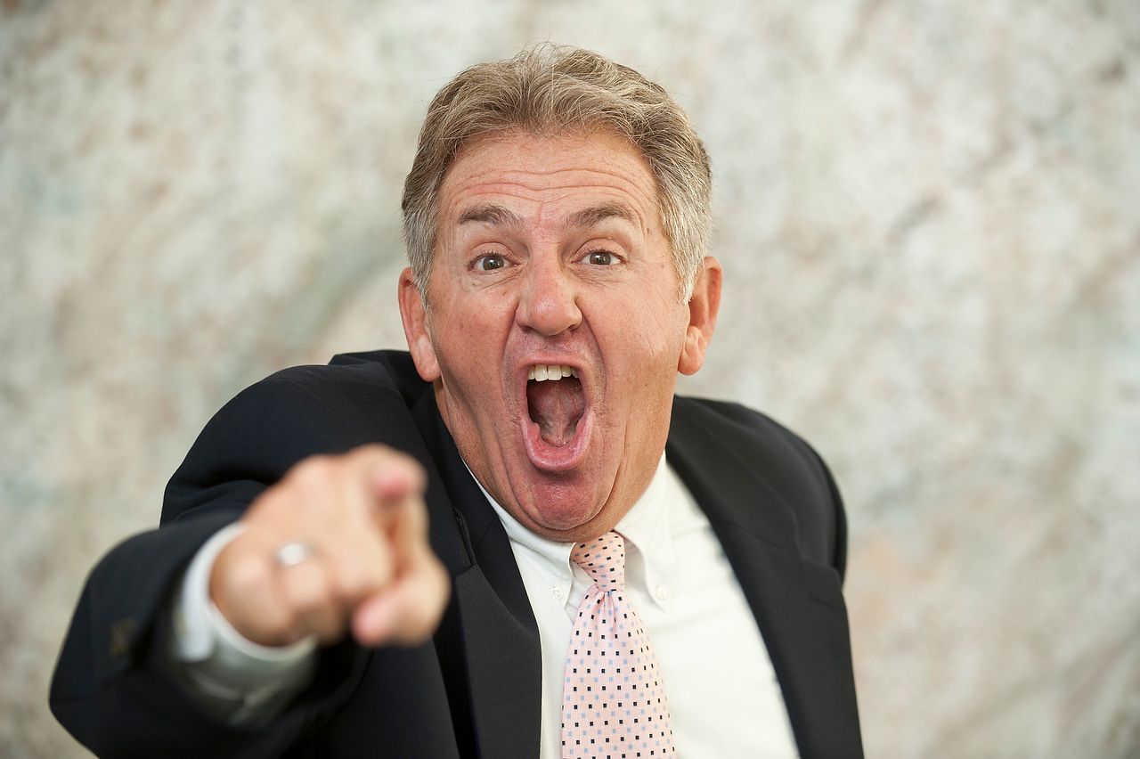 Man in suit angrily pointing, mouth open, against a mottled wall.