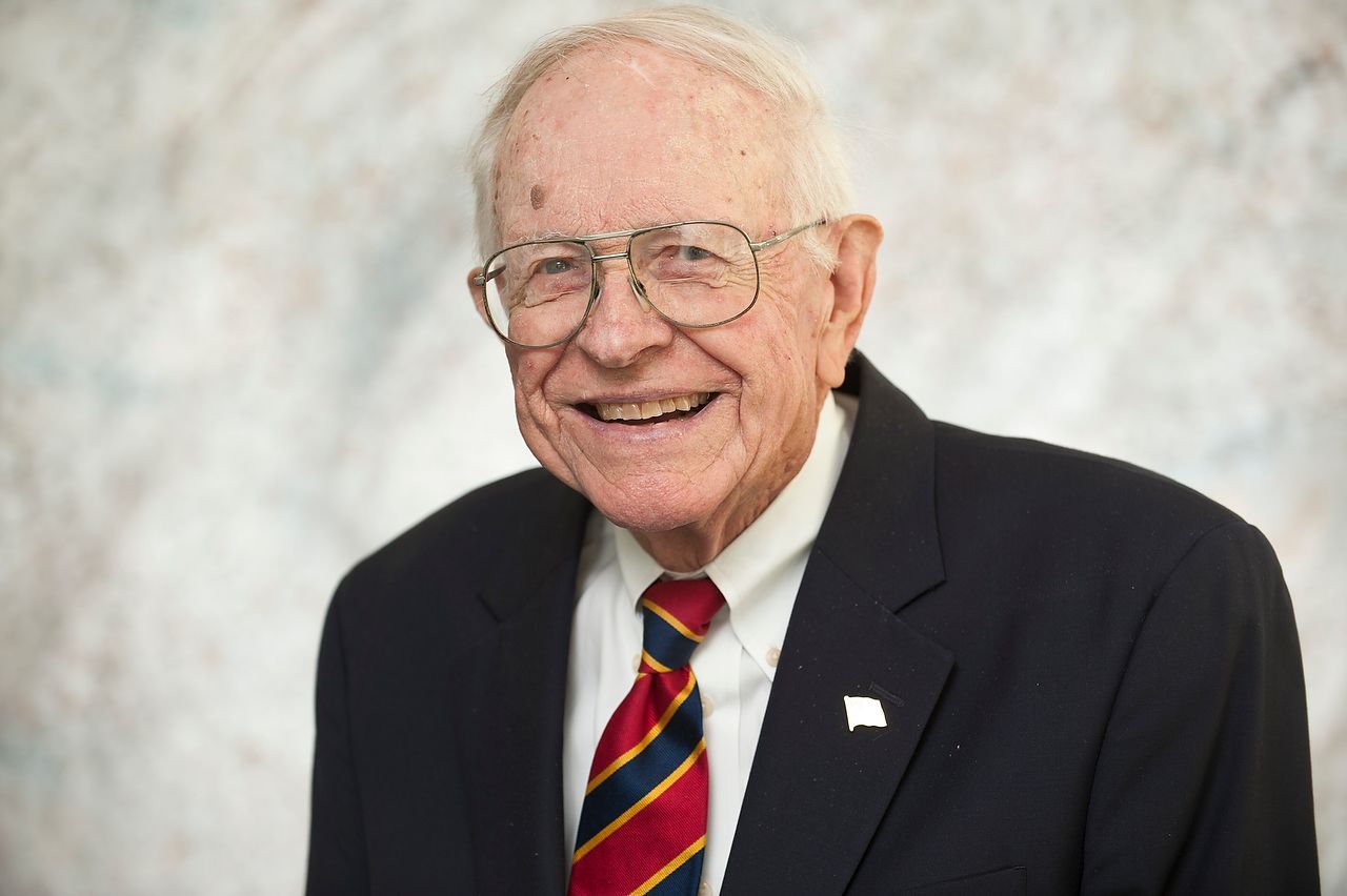 Smiling older man in a suit, tie, and glasses; leans against a light-colored backdrop.