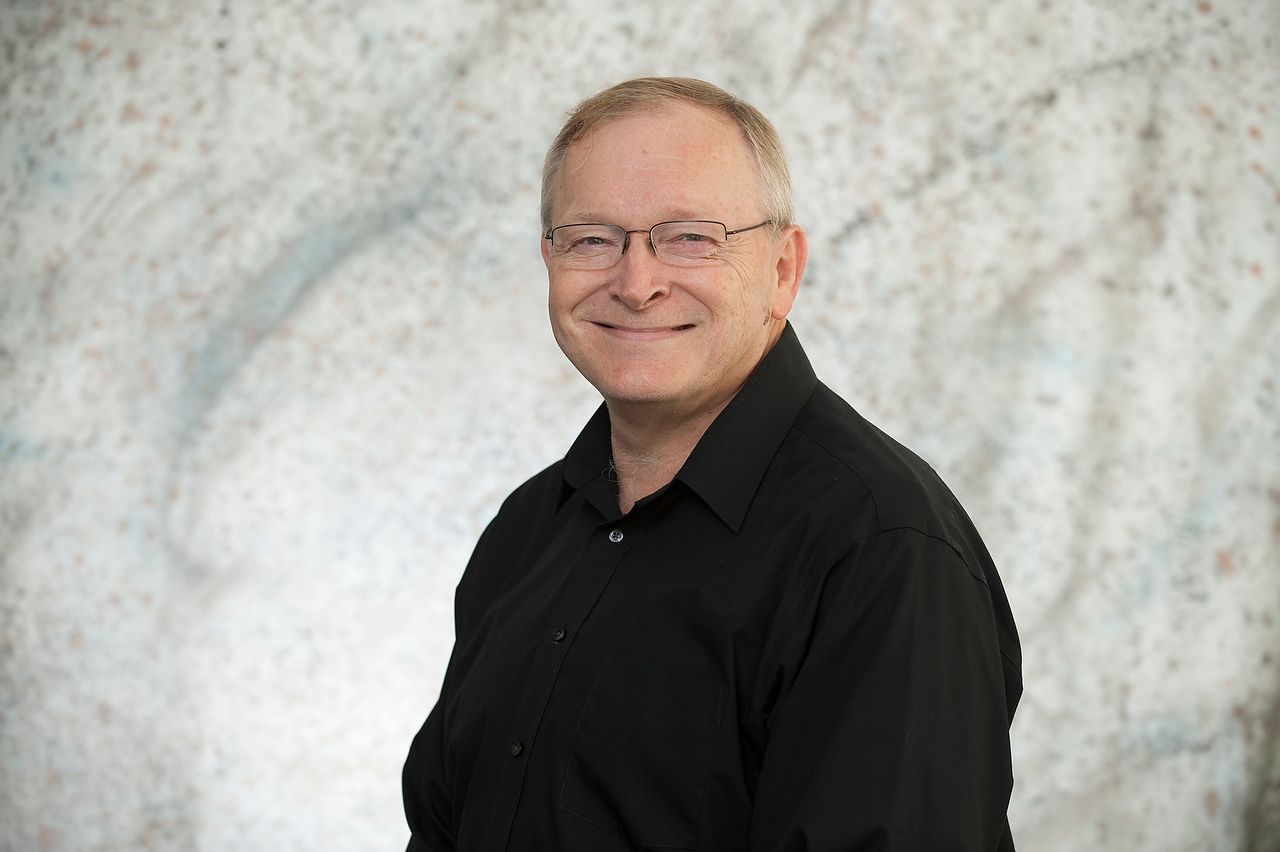 Man with glasses smiles, wearing a black shirt, against a light gray and blue background.