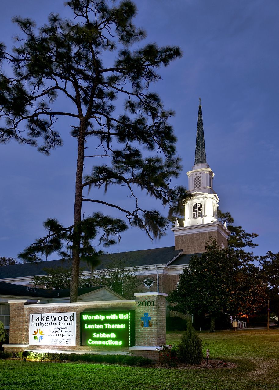 Church at dusk with illuminated sign, tall steeple, and surrounding trees.