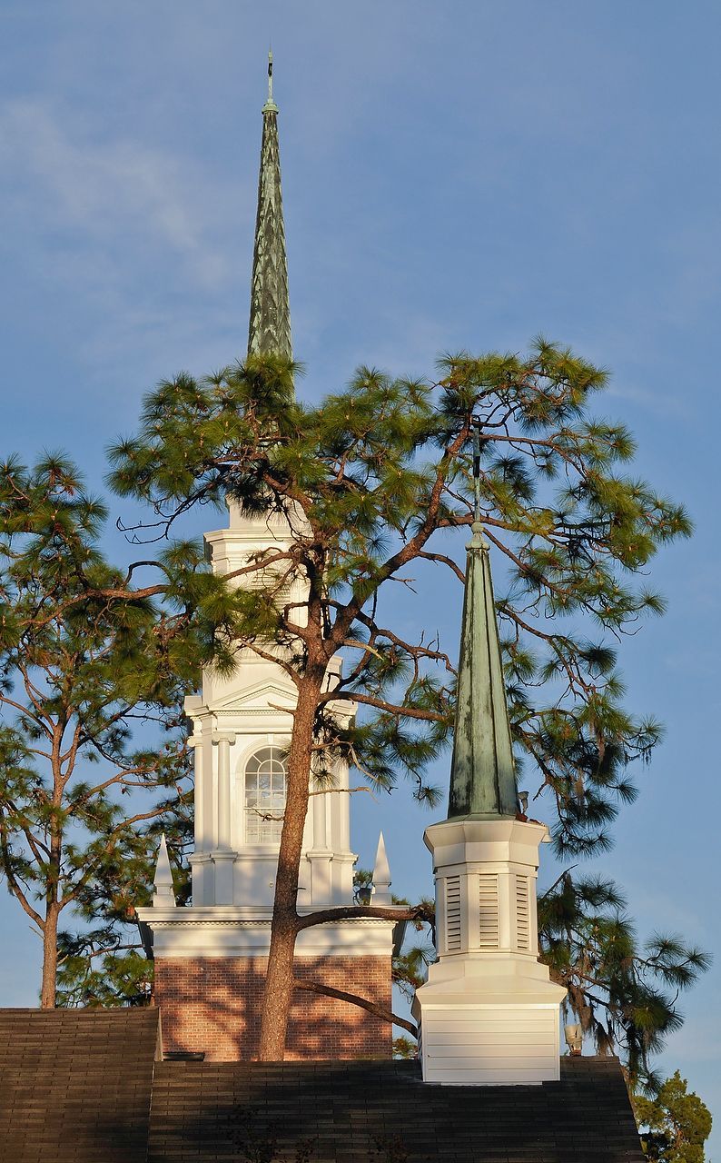 Church steeples rise above trees. White bell tower with pointed spire, green spire to right. Blue sky background.
