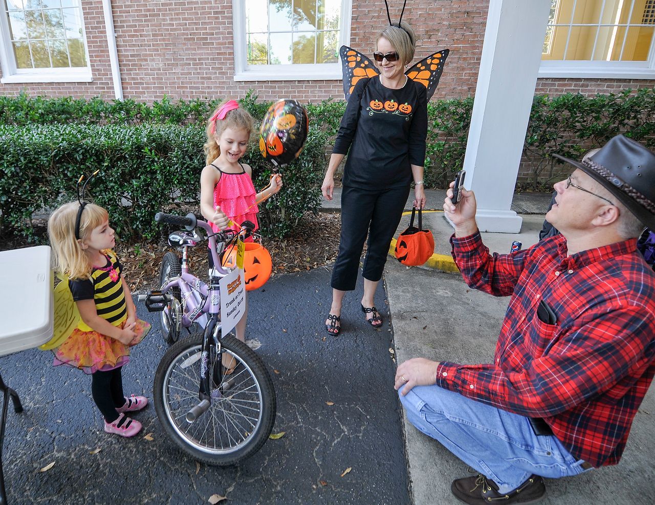Two young children in Halloween costumes, a woman with wings, and a man taking a photo near a bicycle and decorations.