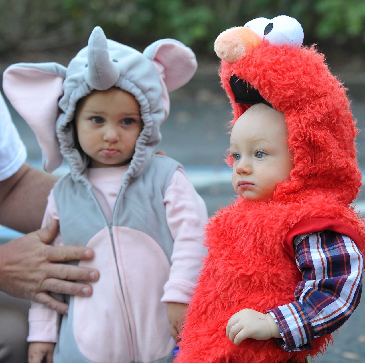 Two babies in costumes: one an elephant, the other Elmo, both looking serious outdoors.