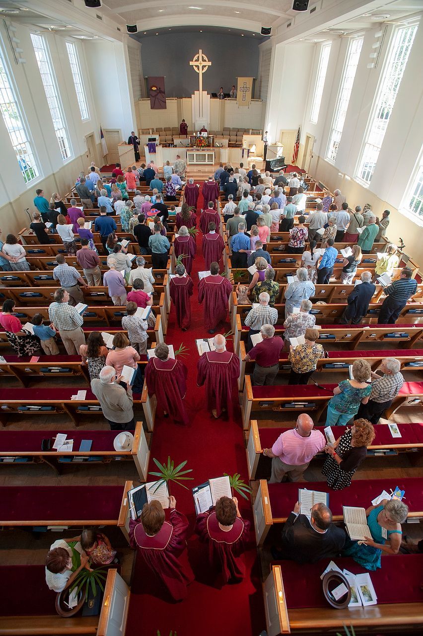 Church service; people in pews, red carpet aisle, clergy in purple robes, cross on altar.
