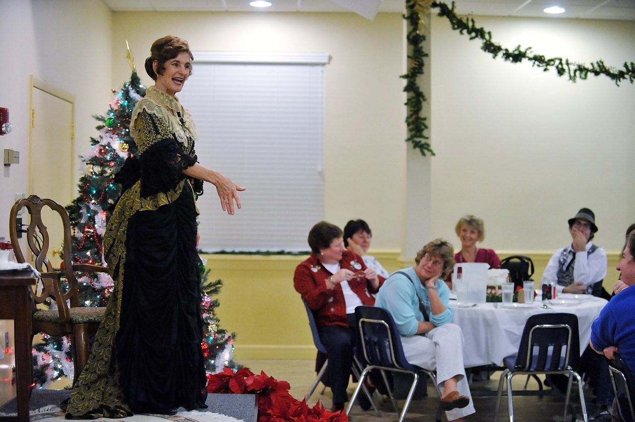 Woman in antique dress performing at a table of people, Christmas tree in the background.
