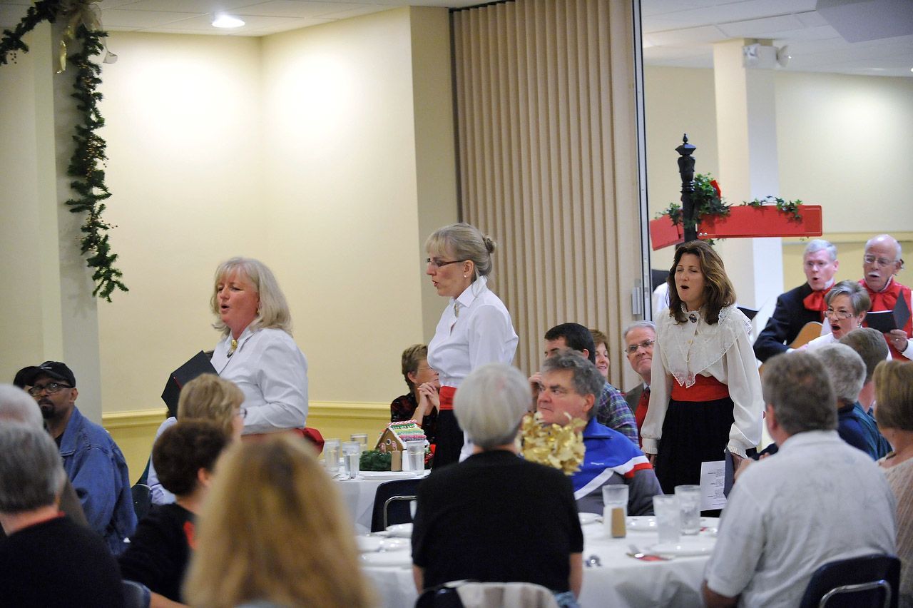People singing in a room with a Christmas decoration. Several seated at tables, some standing, some performing.