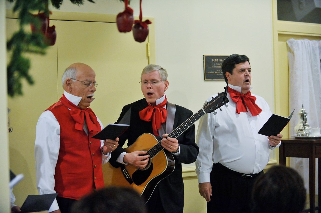 Three men in festive attire sing carols, one with guitar, indoors near Christmas decorations.
