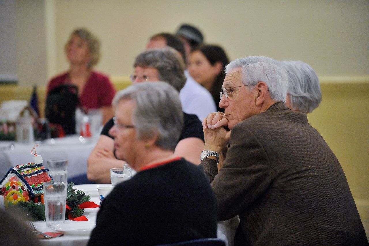 People seated at tables, attentively listening. Some are looking forward, others have their hands on their faces.