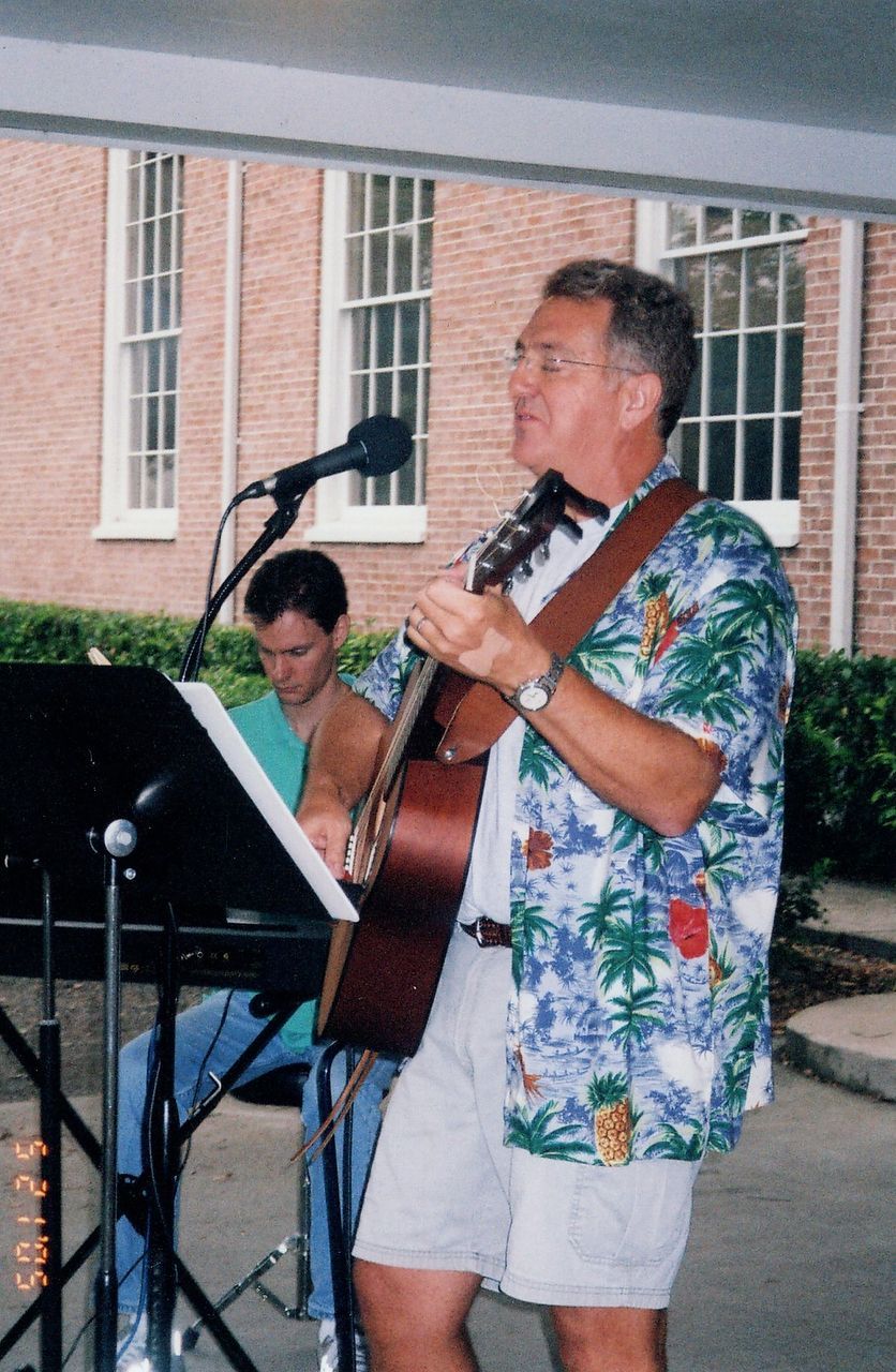 Man singing, playing guitar, wearing a floral shirt, performing outdoors; another person plays keyboard.