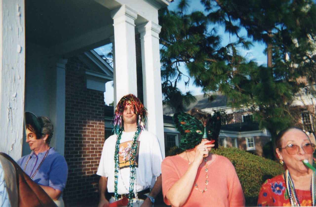 People in festive attire on a porch; one man with a wig and beads. Exterior, brick building with trees.