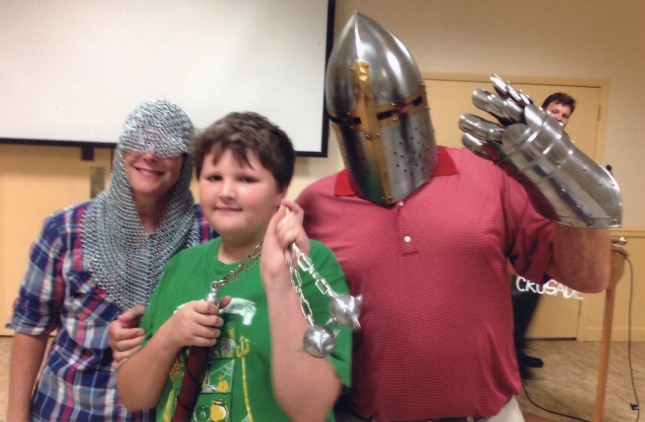 Three people in costume: boy holding flail, one in chainmail, one wearing helmet and gauntlet, indoors.
