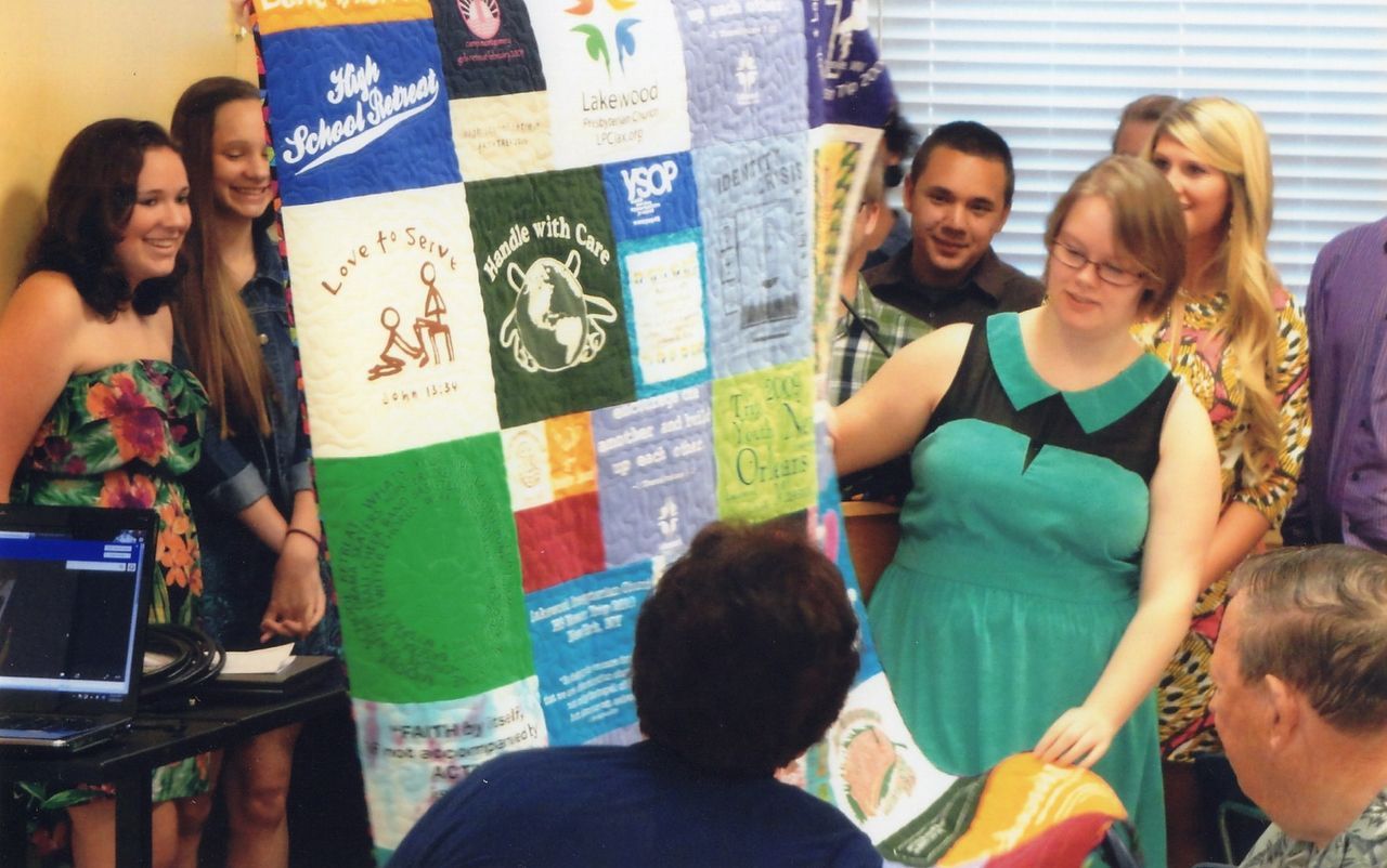 Group admiring a colorful t-shirt quilt. People smile, some point. A laptop sits on a table.