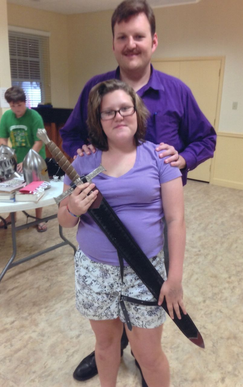 Woman holding sword with man behind her, both wearing purple, in a room.
