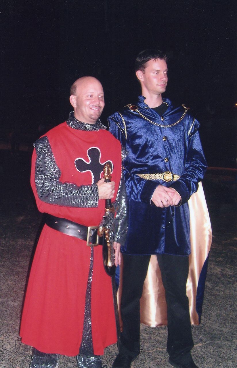 Two men in medieval costumes. One in red with a sword, other in blue with gold trim. Dark outdoor setting.