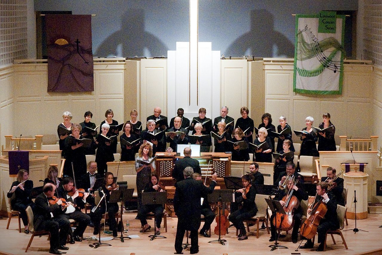 Choir and orchestra performing on stage in a church. Musicians in black attire, banners on the walls.