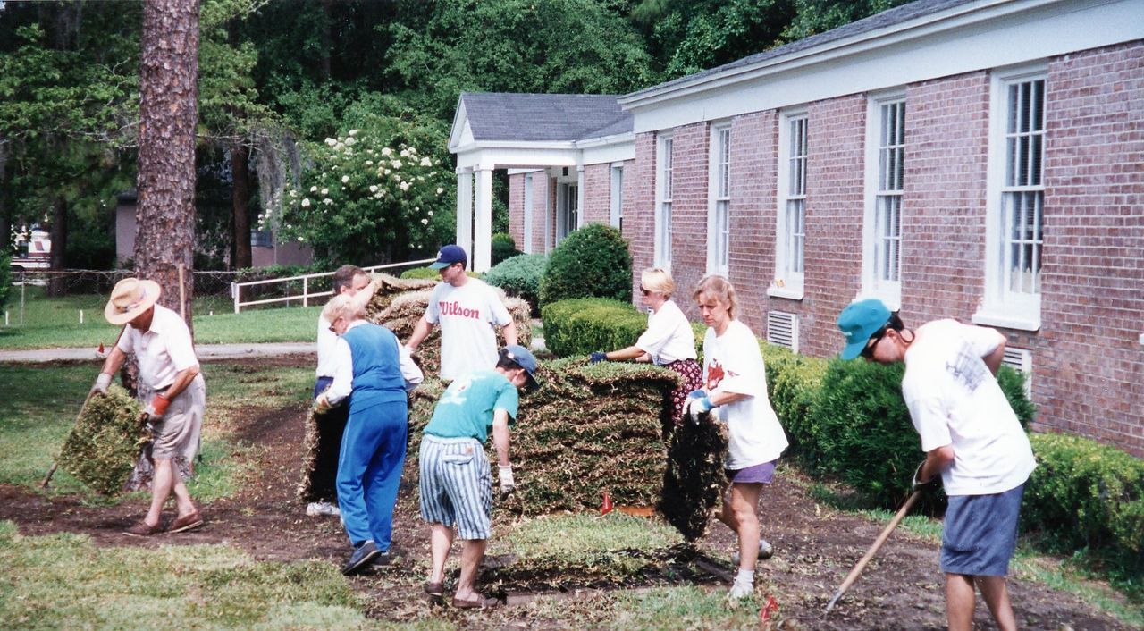 People working in a garden, moving plants near a brick building.