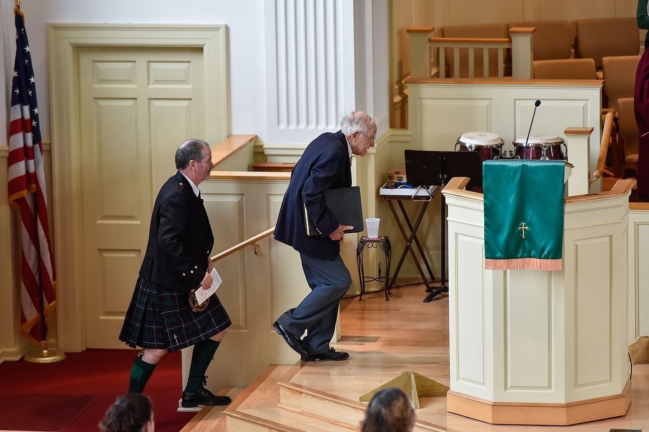 Two men ascend steps into a church. One wears a kilt, the other carries a folder.