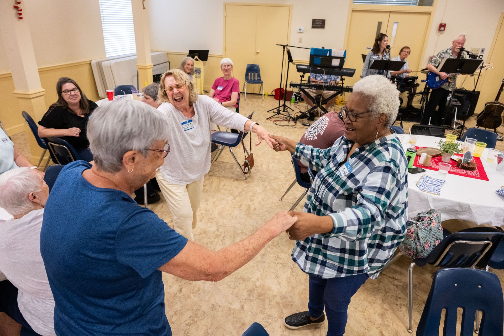 Group of people dancing to a band in a community hall. Joyful expressions, smiling. Band with instruments in the background.