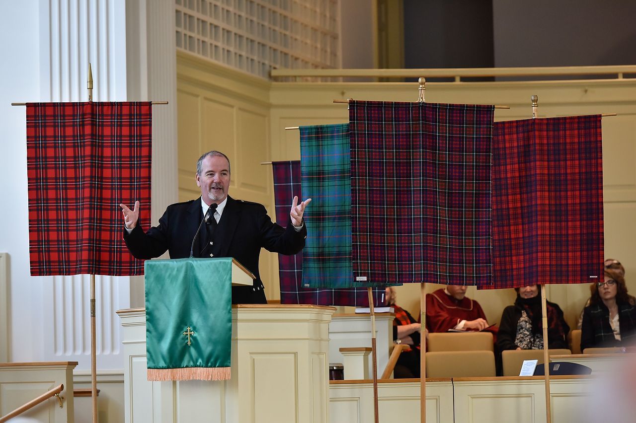 Man speaking at a podium, arms raised, in front of tartan banners. Interior church setting.