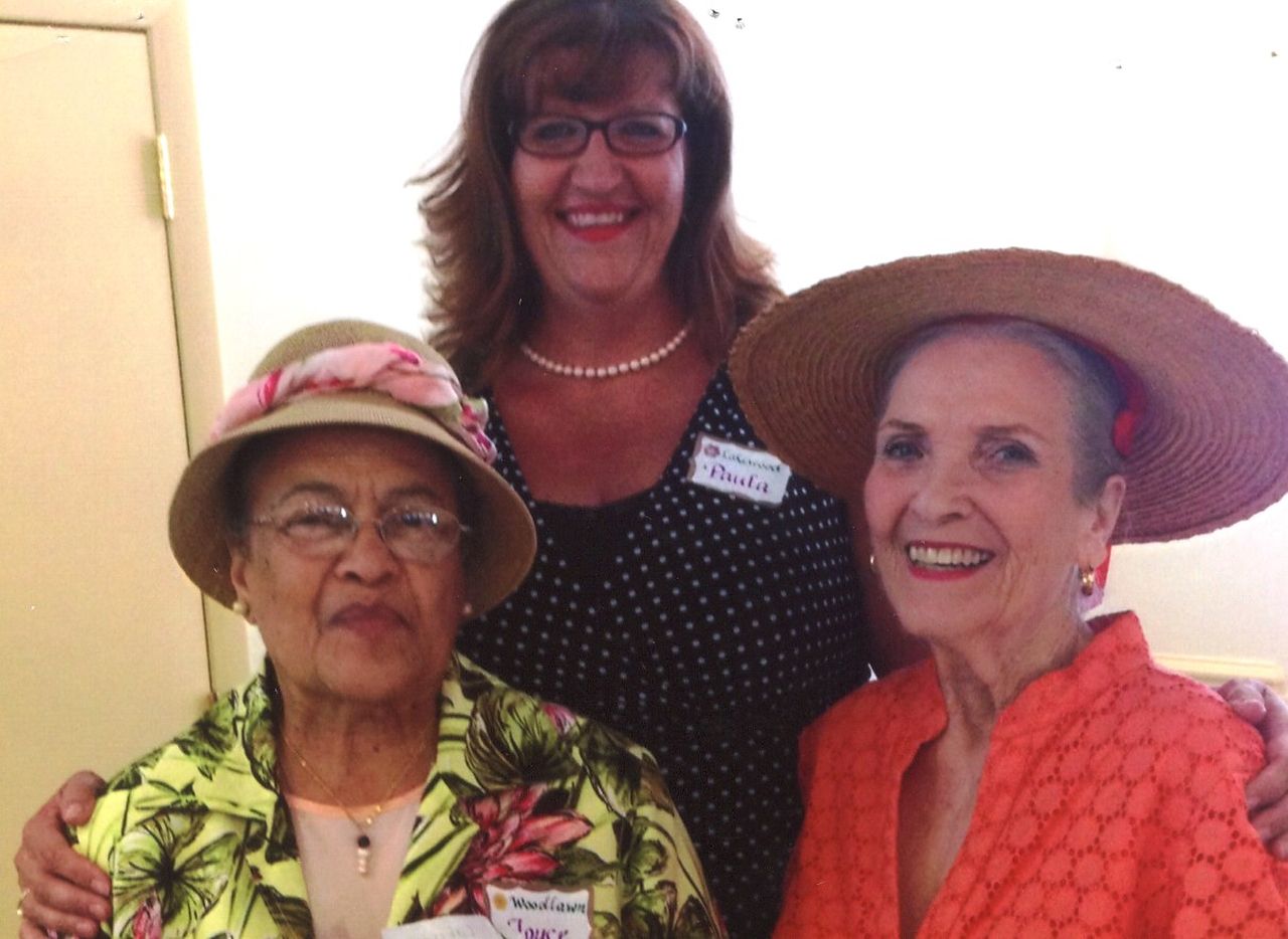 Three women smiling, two in hats and a patterned dress, posing indoors.